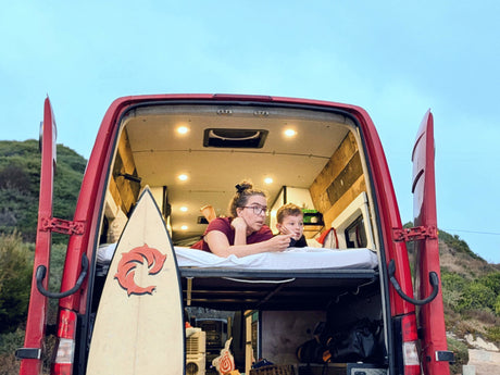 Enjoying van life in an RV rental: A woman and child lounge inside their red camper van, surfboard included. Perfect for family travel.