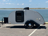 Compact gray teardrop camper RV rental by a serene lake, featuring a distinctive door and black accents on a sunny day.