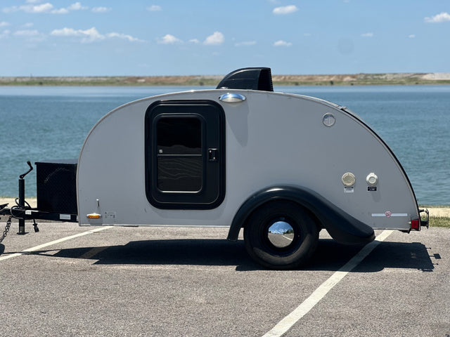 Compact gray teardrop camper RV rental by a serene lake, featuring a distinctive door and black accents on a sunny day.
