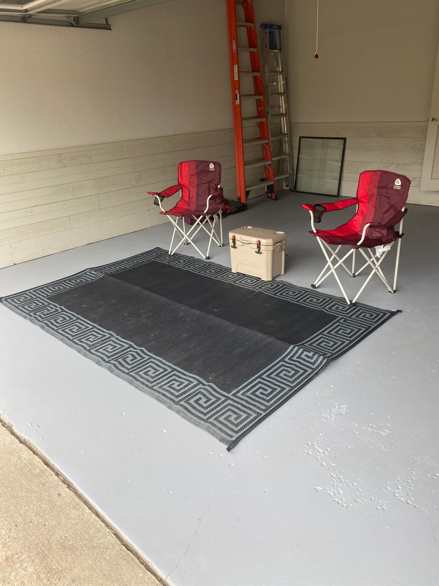 RV rental trip essentials: Two red camping chairs, a tan cooler, and a patterned rug laid out on a light grey garage floor.