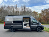 Dark gray camper van, ready for your RV rental adventure. Side door open reveals a compact interior. Parked on asphalt near trees.