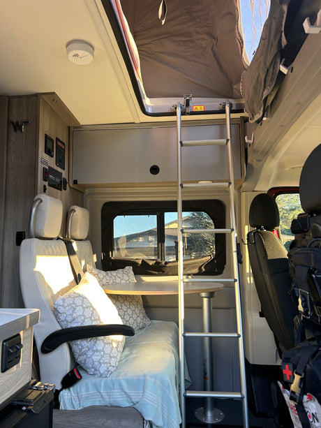 Bright RV rental interior: Ladder to pop-top bed, light-colored seating, and dining table. A cozy camper van.
