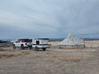 Adventure-ready RV rental: White Tacoma tows an overland trailer past art-covered tents in a dusty desert landscape.