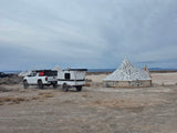 Adventure-ready RV rental: White Tacoma tows an overland trailer past art-covered tents in a dusty desert landscape.