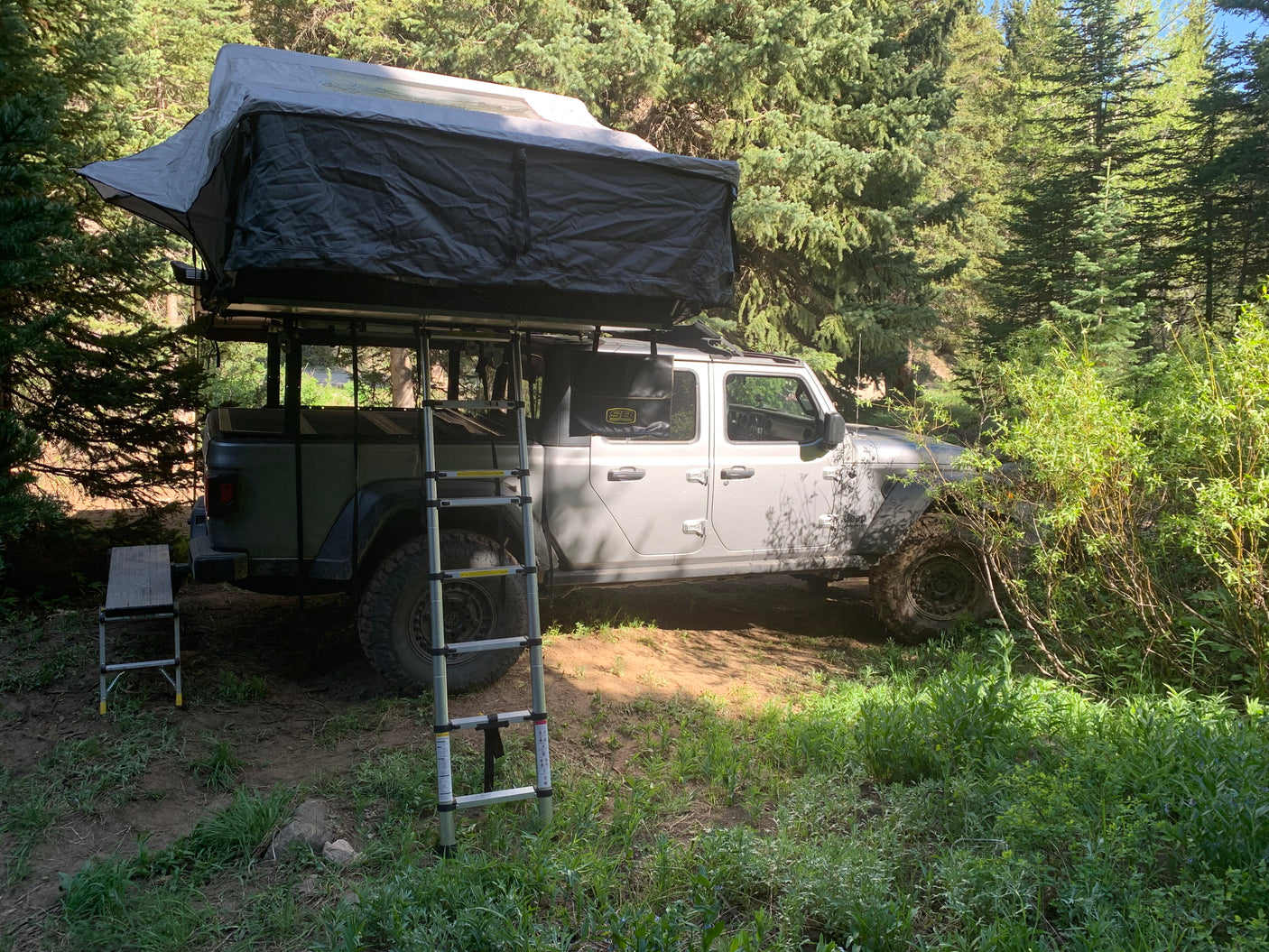 Grey Jeep Gladiator with roof-top tent set up for camping in a lush forest campsite. A modern rv rental adventure option.