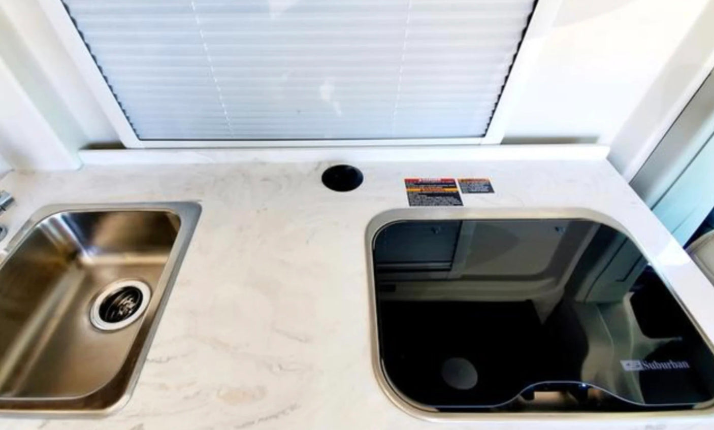 RV rental kitchen featuring a stainless steel sink, covered glass cooktop, and light countertop under a window with blinds.