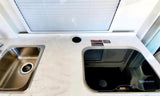 RV rental kitchen featuring a stainless steel sink, covered glass cooktop, and light countertop under a window with blinds.