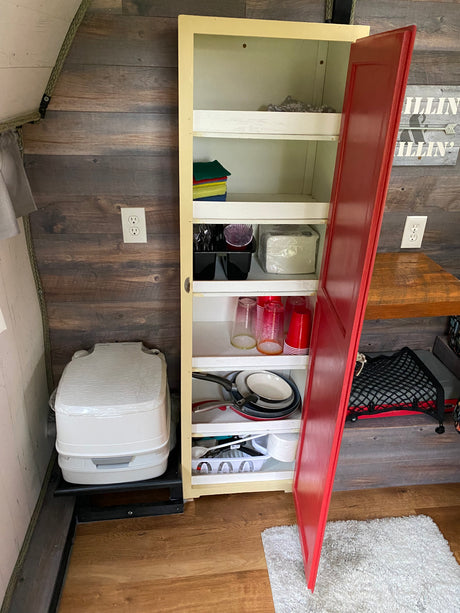RV rental organized storage: Cabinet with kitchen supplies, pans, cups, and a portable toilet for camping convenience.