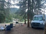 RV rental van at a forest campsite. A person tends a campfire by a river, surrounded by tall pine trees and mountains.