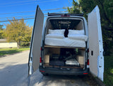Open rear doors of a white RV rental reveal a comfortable elevated bed and organized storage space below for gear.