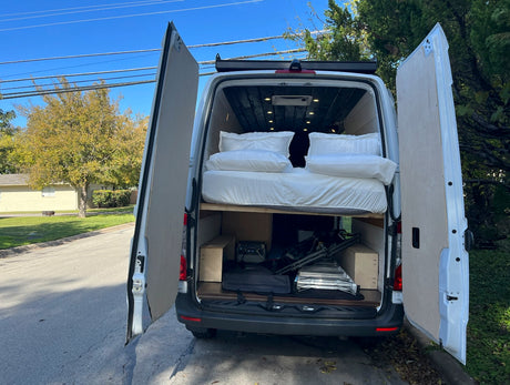 Open rear doors of a white RV rental reveal a comfortable elevated bed and organized storage space below for gear.