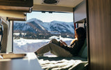 A woman relaxes in her rv rental, reading a book with a snowy mountain view from her cozy, sunlit interior. Winter escape.