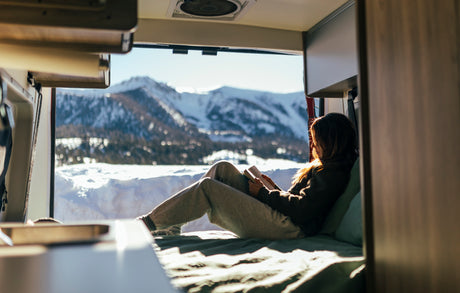 A woman relaxes in her rv rental, reading a book with a snowy mountain view from her cozy, sunlit interior. Winter escape.