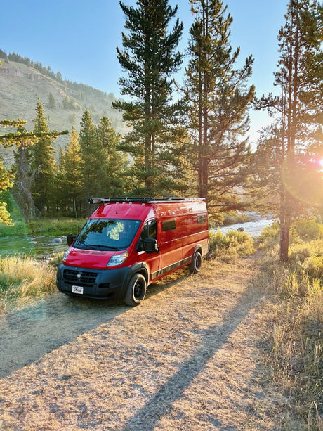 Red RV rental parked by a river, nestled among sunlit trees & mountains on a dirt path. Your perfect outdoor escape awaits.