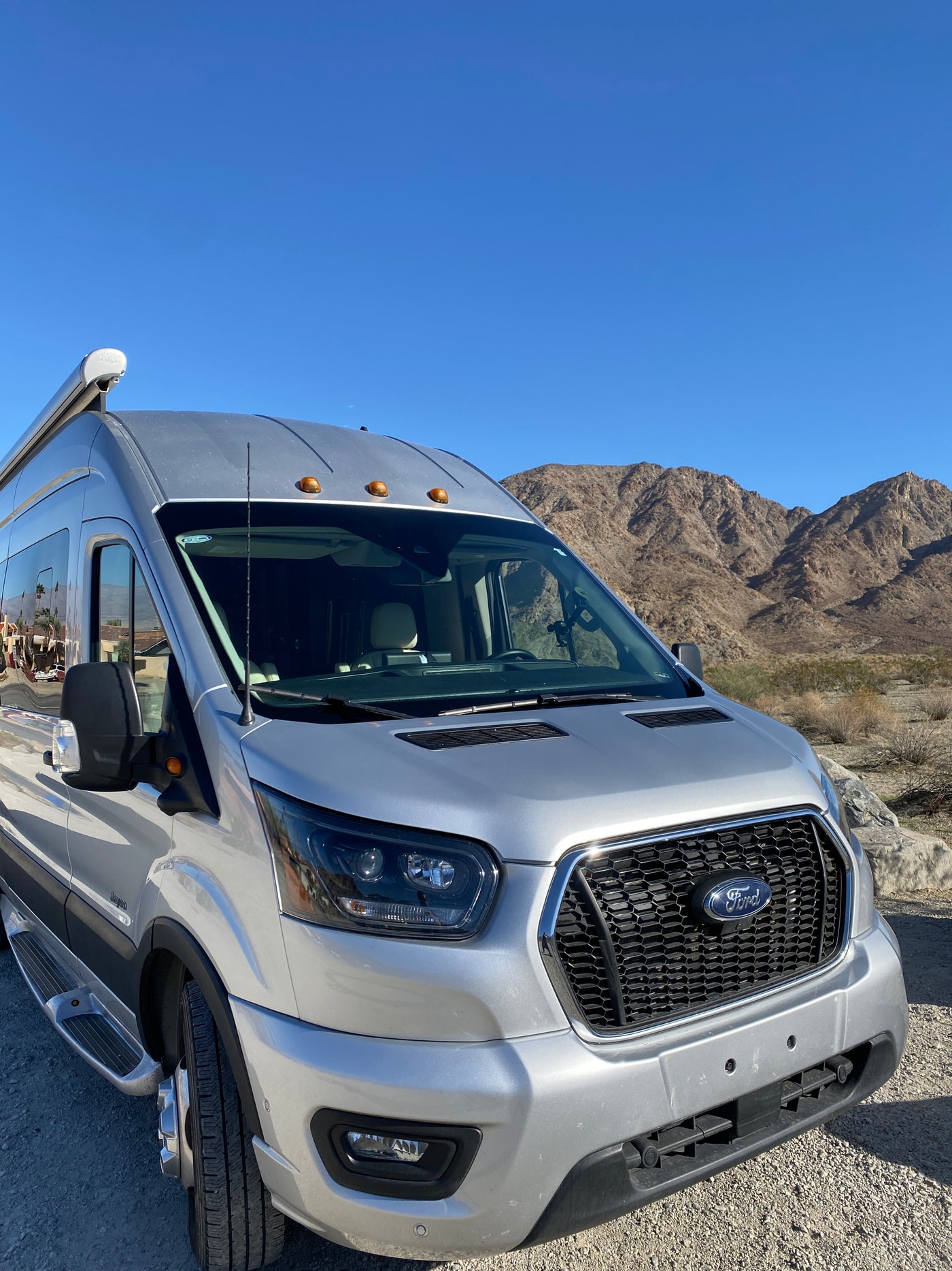 Silver Ford Transit RV rental parked in front of desert mountains under a clear blue sky, ready for adventure. — photo 7