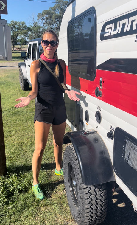 Woman presents a red and white "SUNRAY" RV rental with a white Jeep in the background, ready for adventure.