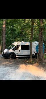 White camper van RV rental with blue logo parked at a wooded campsite, ready for exploration.