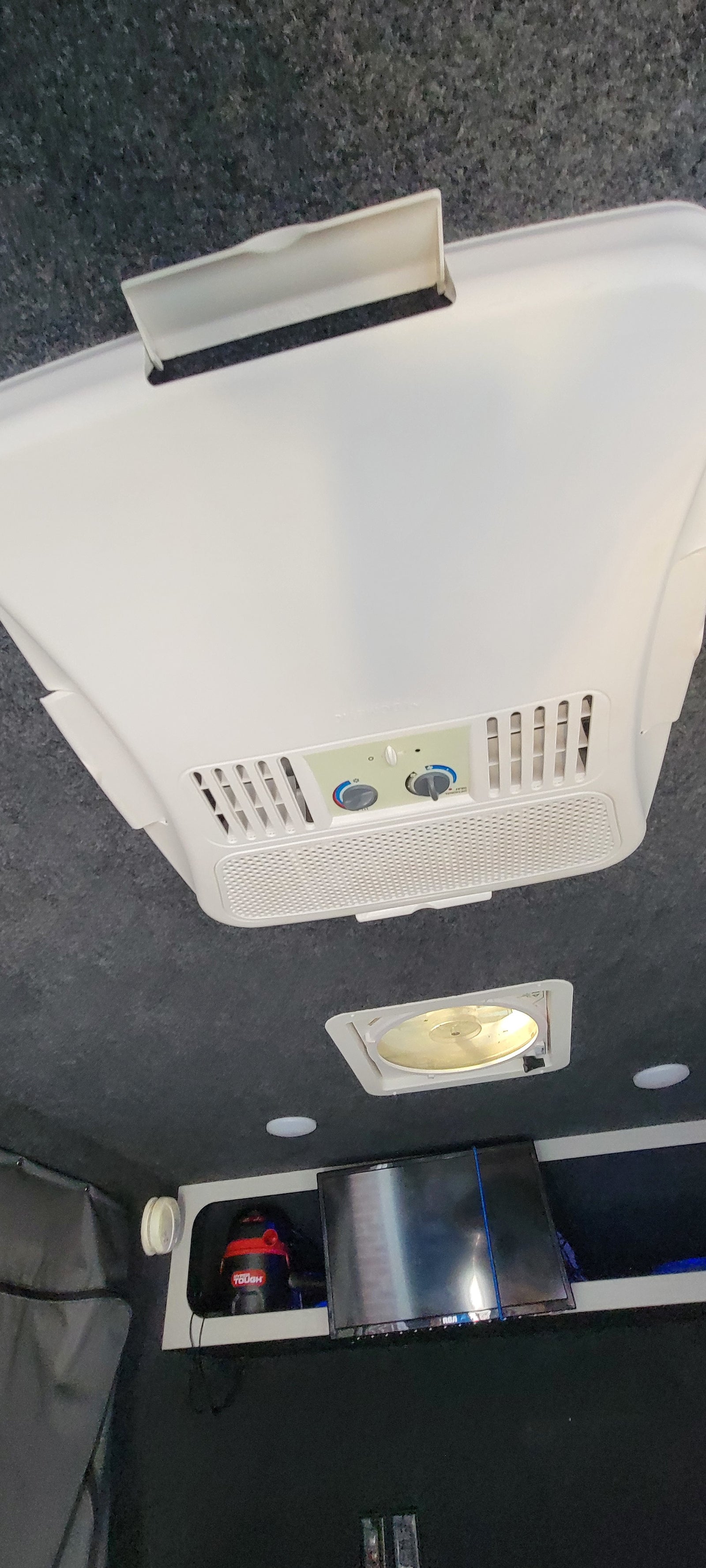 Inside an RV rental: White ceiling-mounted AC unit, roof vent, TV, and vacuum cleaner. Equipped for travel comfort.