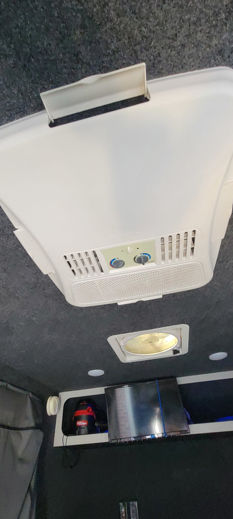 Inside an RV rental: White ceiling-mounted AC unit, roof vent, TV, and vacuum cleaner. Equipped for travel comfort.