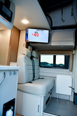 Inside an RV rental, a modern camper van lounge features a TV displaying "Game Plan Age +40", plush seating, and a stowed table.