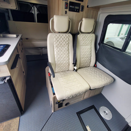View of a comfortable RV rental interior with beige quilted seats, a compact kitchen counter, and control panel. Textured floor.
