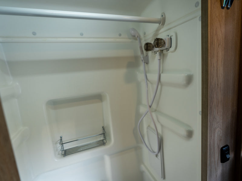 Compact white RV rental shower stall featuring a handheld shower, water controls, and a wire shelf. — photo 12