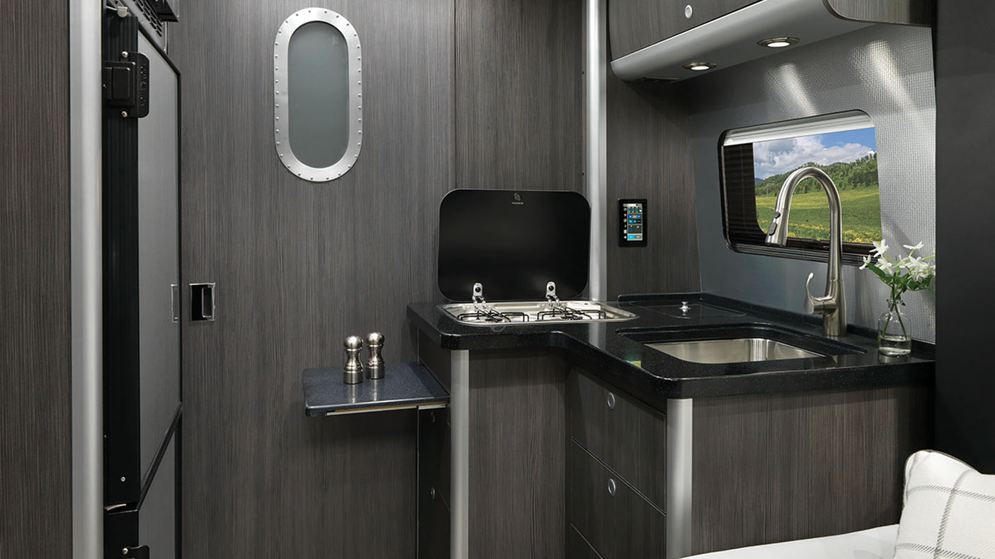 Modern RV rental interior showing a compact kitchen with a sink, stove, and dark wood cabinetry.