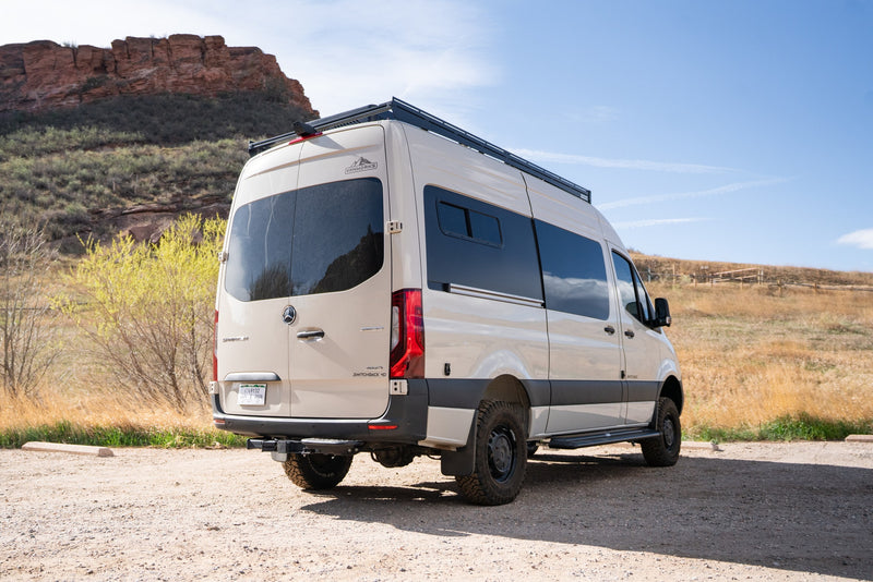 Tan Mercedes Sprinter adventure van for rv rental, parked on a gravel path by mountains and bushes under a clear sky. — photo 2