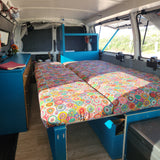 Cozy RV rental interior with vibrant beds, blue storage, and views of nature for comfortable travel.