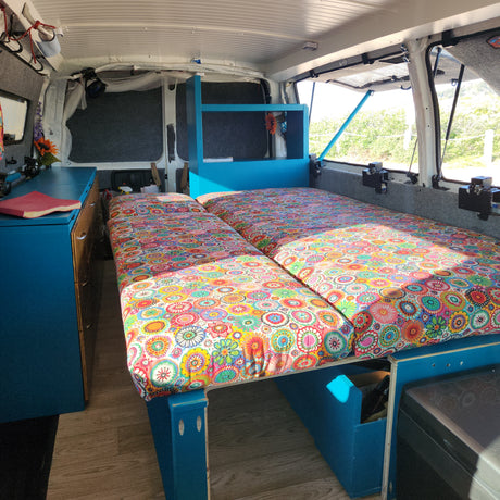 Cozy RV rental interior with vibrant beds, blue storage, and views of nature for comfortable travel.