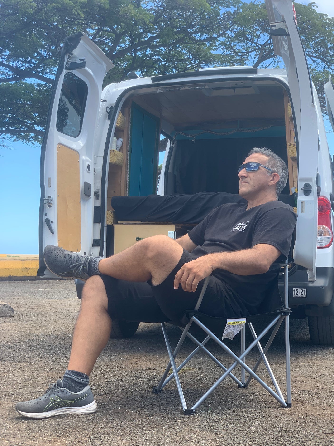 Man relaxing on a chair outside an RV rental with an open rear hatch showing its customized camper interior.