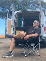 Man relaxing on a chair outside an RV rental with an open rear hatch showing its customized camper interior.