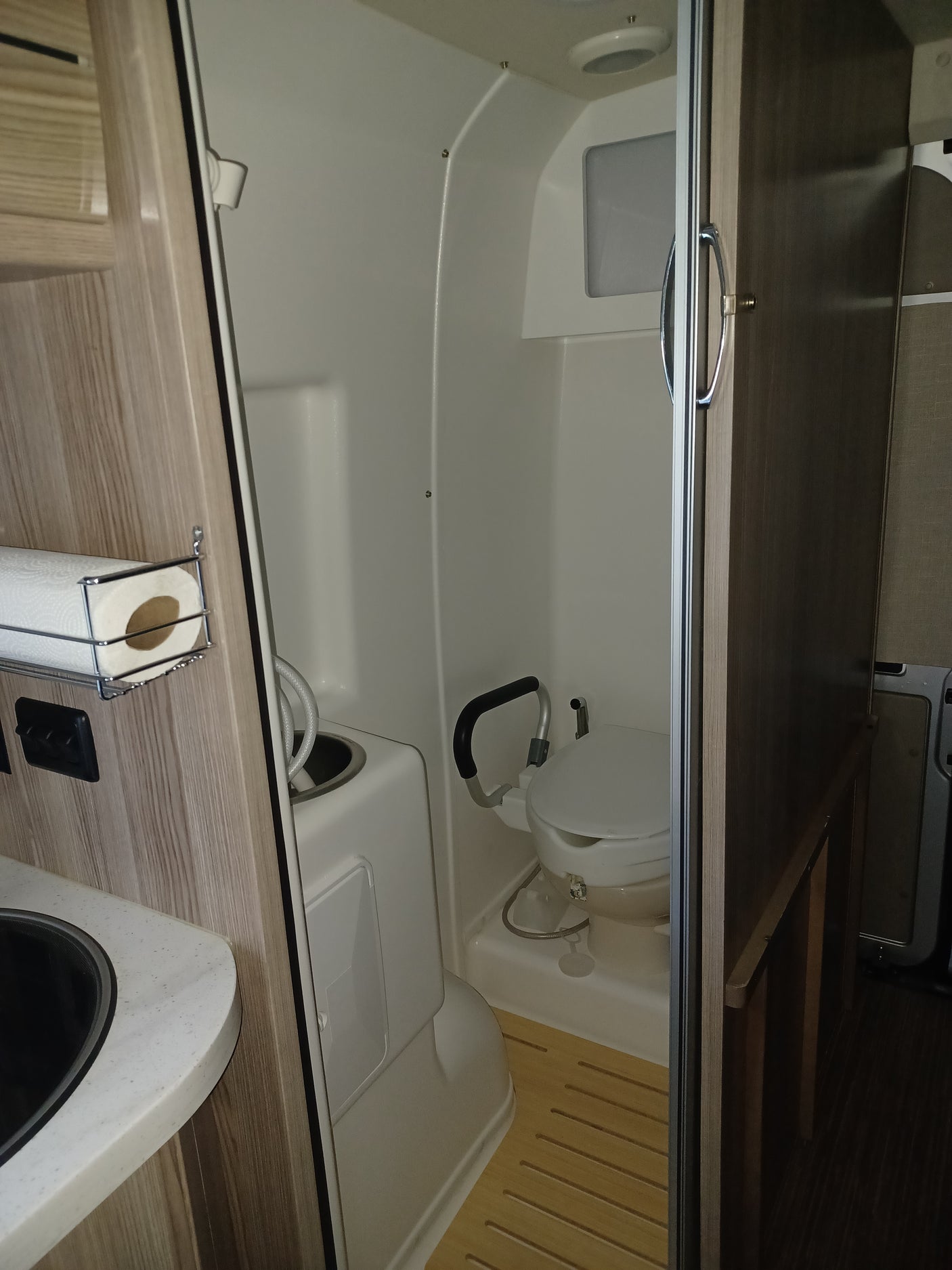 Compact rv rental wet bath interior showing a white toilet, shower hose, slatted wooden floor, and a paper towel holder.