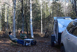 Two people relax in camping chairs by an RV rental and car in a peaceful forest setting.
