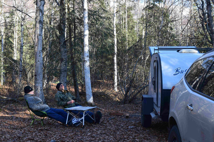 Two people relax in camping chairs by an RV rental and car in a peaceful forest setting.