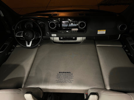 RV rental camper van interior: front seats converted to a bed, Mercedes-Benz dashboard, and steering wheel visible.