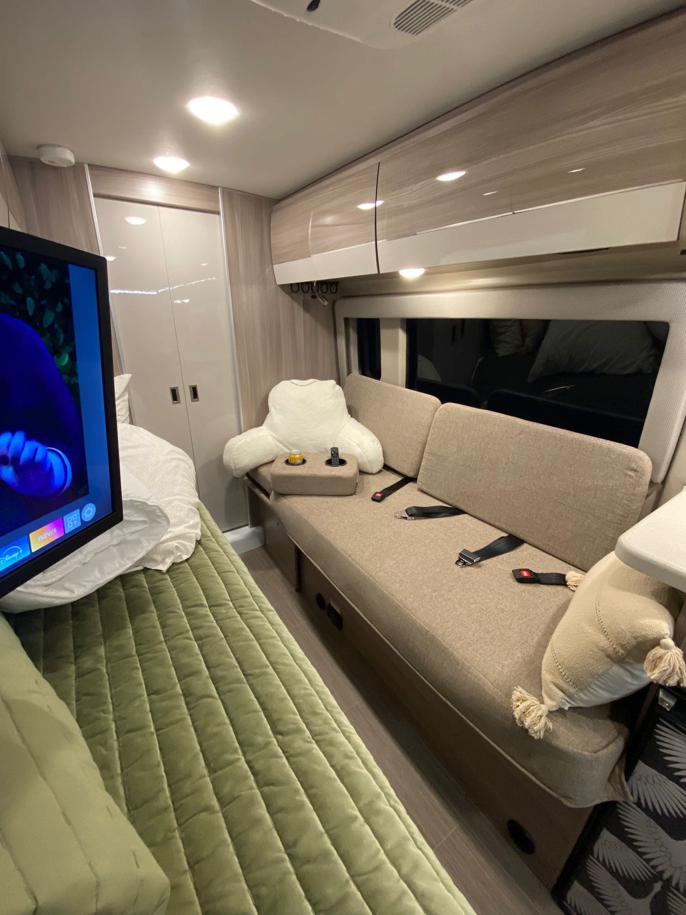 RV rental interior featuring a cozy green bed, TV, and convertible bench seating with seatbelts.