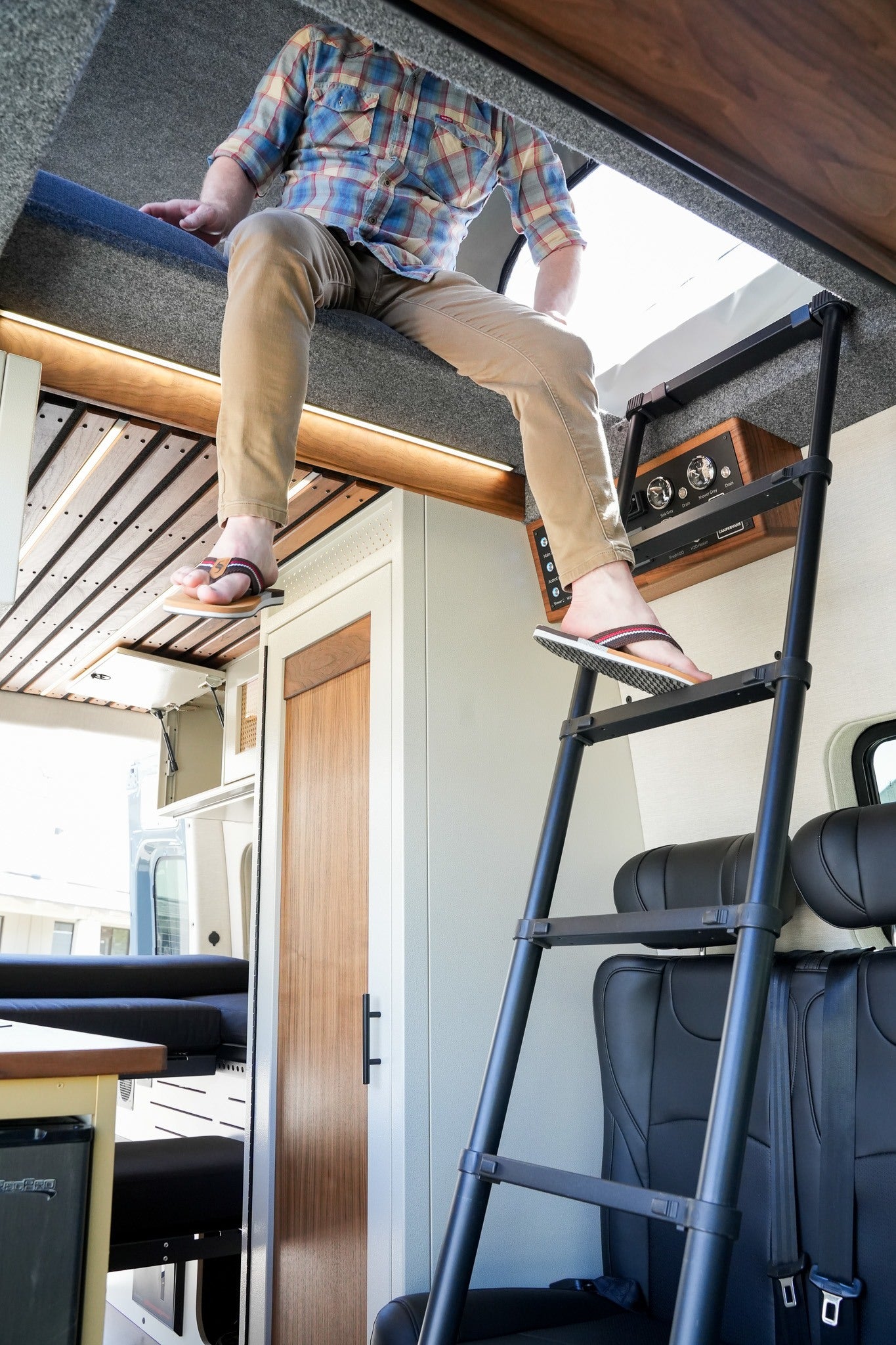 Person on RV rental's top bunk, wearing flip-flops, about to use the ladder in this compact camper van interior.
