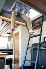 Person on RV rental's top bunk, wearing flip-flops, about to use the ladder in this compact camper van interior.