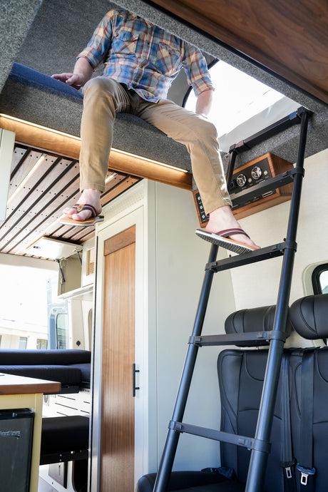 Person on RV rental's top bunk, wearing flip-flops, about to use the ladder in this compact camper van interior.