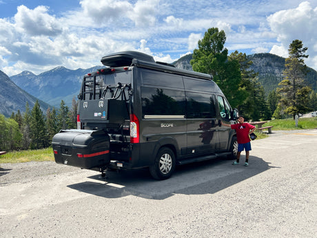 A dark gray camper van, an RV rental, with a boy posing in front of it. Majestic mountains and a cloudy sky form the backdrop.