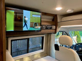 An RV rental's compact interior features a wall-mounted TV, stereo, and large window overlooking a fence. Bright and cozy.