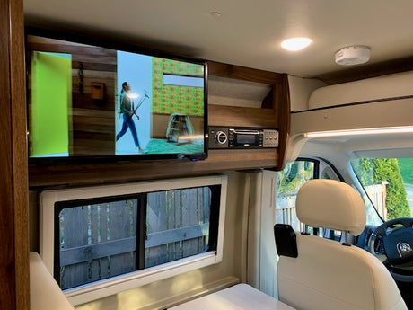 An RV rental's compact interior features a wall-mounted TV, stereo, and large window overlooking a fence. Bright and cozy.
