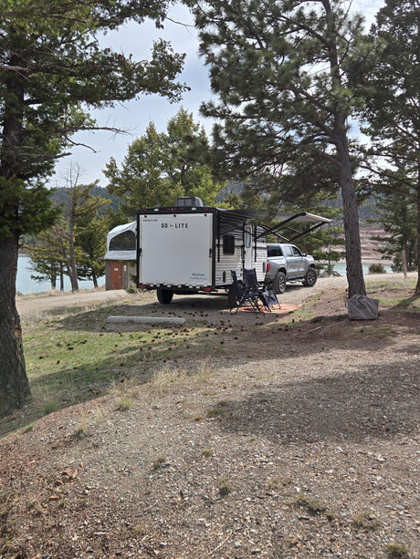 Lakeside rv rental setup with a white travel trailer, truck, and camping chairs under pine trees by the water.