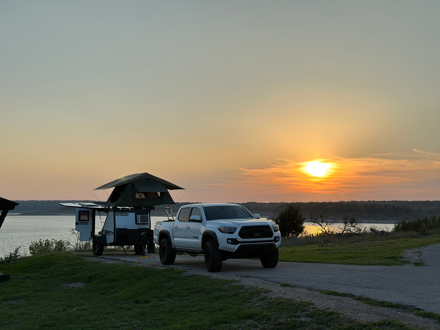 Stunning sunset over a lake with a white Toyota Tacoma and a TigerMoth overland trailer with a rooftop tent, ideal for rv rental adventures.