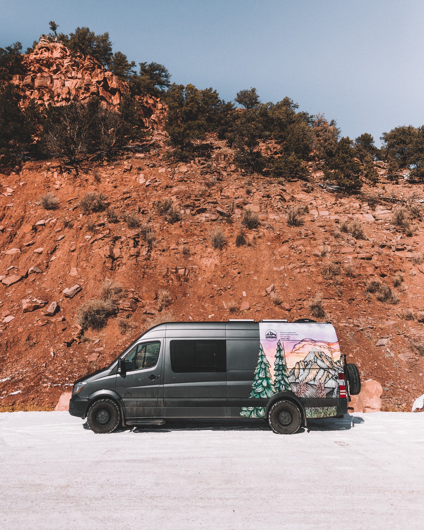 Dark grey RV rental van with custom mountain art, parked on a snowy road against a towering red rock cliff under a blue sky.