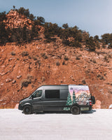 Dark grey RV rental van with custom mountain art, parked on a snowy road against a towering red rock cliff under a blue sky.