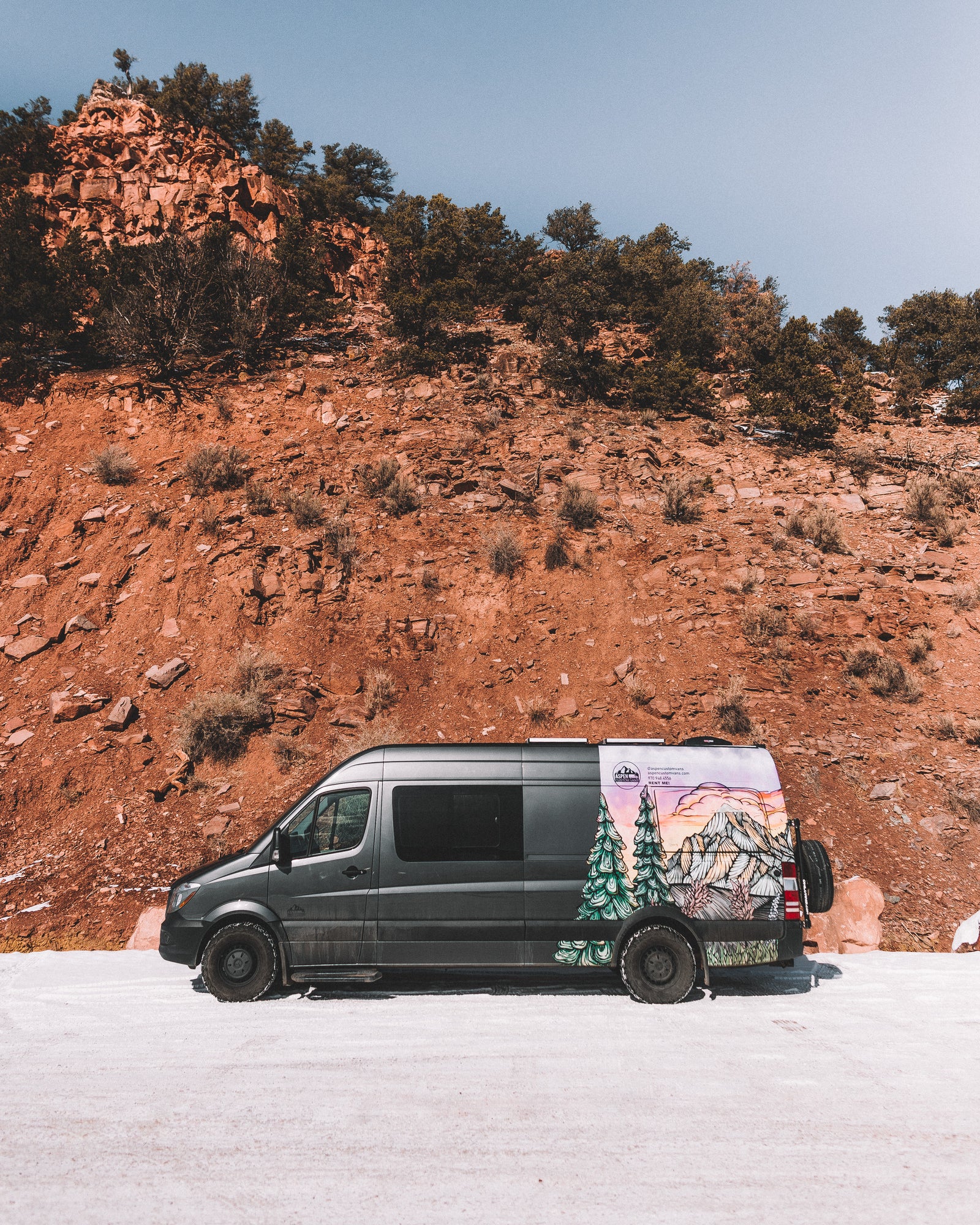 Dark grey RV rental van with custom mountain art, parked on a snowy road against a towering red rock cliff under a blue sky.