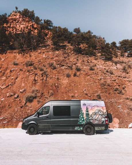 Dark grey RV rental van with custom mountain art, parked on a snowy road against a towering red rock cliff under a blue sky.