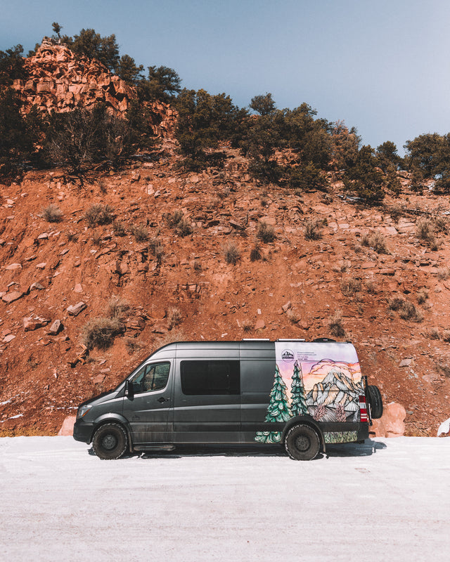 Dark grey RV rental van with custom mountain art, parked on a snowy road against a towering red rock cliff under a blue sky.